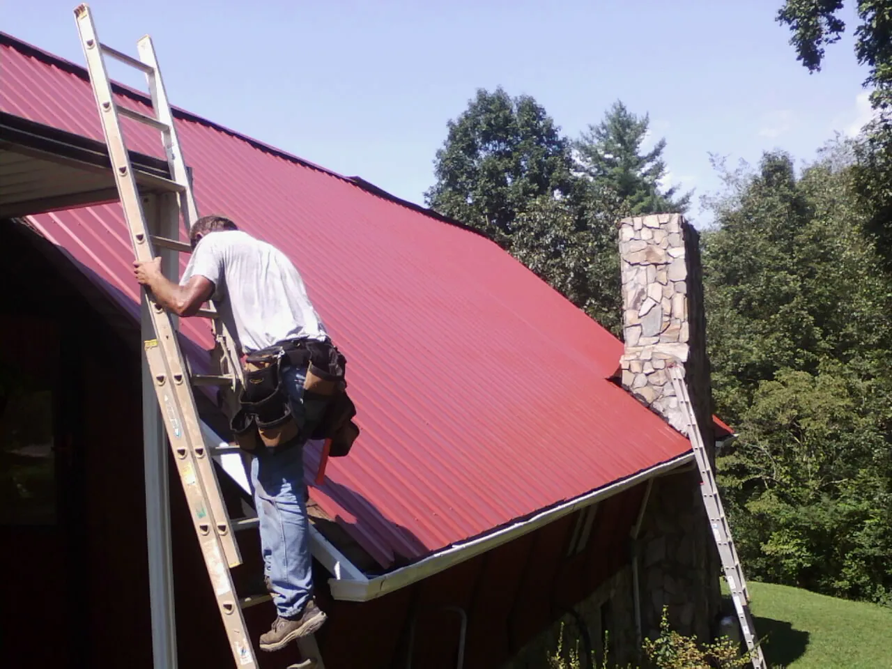 Roofer on a ladder beside a red metal roof and stone chimney — Soffit & Fascia Repair by Roofing Brothers in Crozet